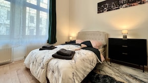 Un dormitorio con una cama, dos mesitas de noche, un tocador, una ventana con cortinas y un cuadro de un paisaje urbano en la pared.