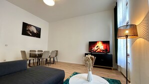 Una sala moderna con un sofá, una mesa de comedor con sillas, un televisor que muestra una chimenea y una lámpara de pie.