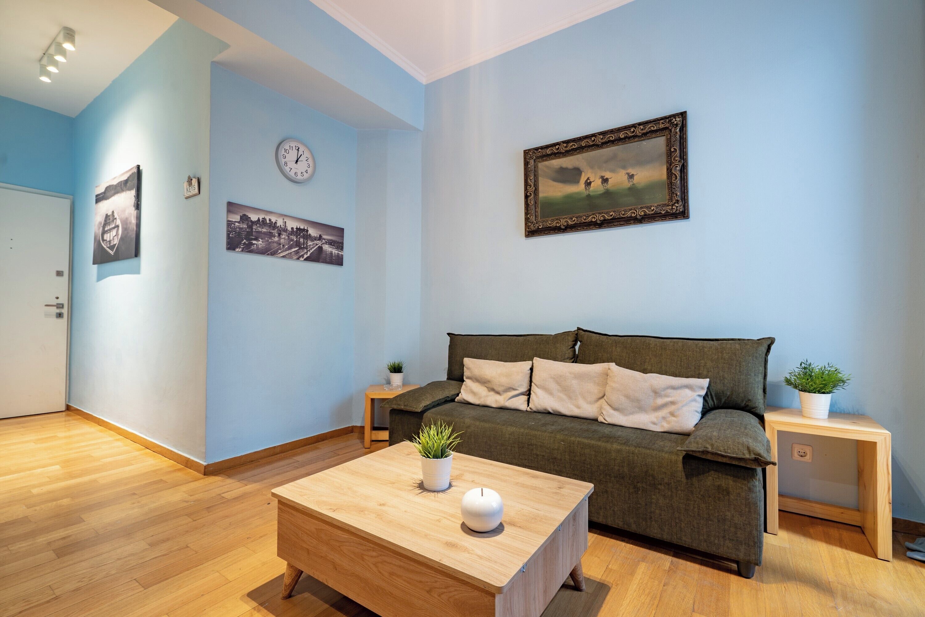 Een woonkamer met een bank, een bijzettafeltje met een plant, een klok en foto's in lijsten aan de muur.
