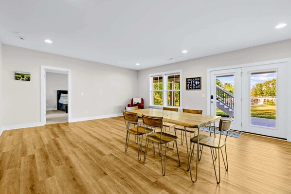 Una habitación amplia con una gran mesa de madera y sillas, paredes blancas y ventanas grandes que ofrecen vistas a un paisaje verde.