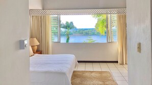 A bedroom with a large window, a bed, a lamp, and a view of a lake and greenery.