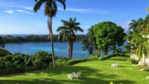 A lush green lawn with white chairs, palm trees, and a view of a calm sea.