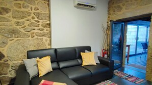 A living room with a black leather sofa, two yellow cushions, a red and yellow checkered throw blanket, a colorful woven rug, and a stone wall. - Casa do Parada | Private Pool | Rural Refuge (Messegães Monção)