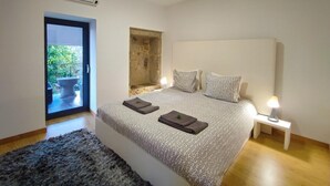 A bedroom with a large bed, a small bedside table with a lamp, a stone wall, and a view of greenery outside. - Casa do Parada | Private Pool | Rural Refuge (Messegães Monção)