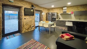 A modern kitchen with stone walls, a dining area with white chairs, and a TV mounted on the wall. - Casa do Parada | Private Pool | Rural Refuge (Messegães Monção)