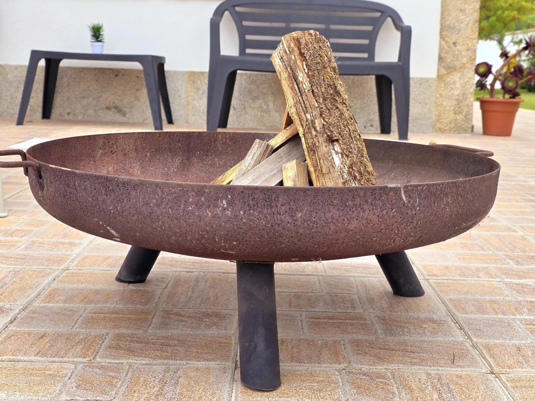 Un foyer métallique contenant du bois, placé sur une terrasse carrelée, avec des chaises et une petite table en arrière-plan.