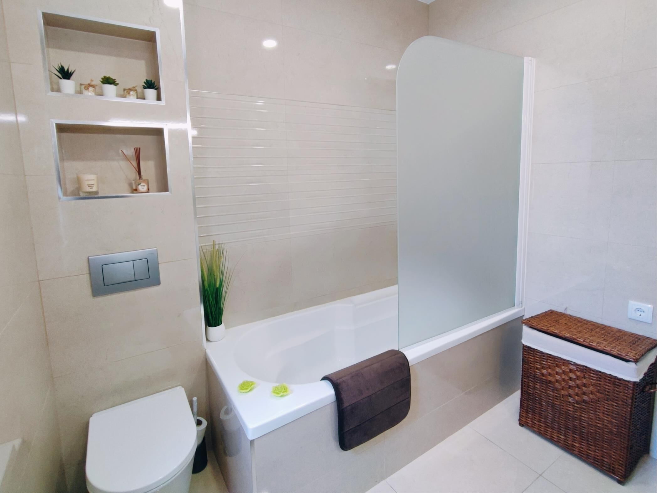 A modern bathroom with a toilet, a shower, and a sink. There are shelves with decorative items and a small plant.