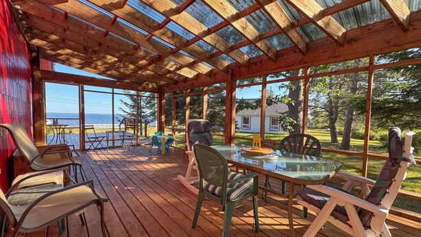 Un espace extérieur aménagé, partiellement abrité, avec une terrasse en bois, une table en verre et des chaises, offrant une vue sur un plan d’eau et une zone herbeuse bordée d’arbres.