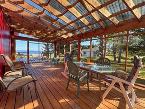 Un espace extérieur aménagé, partiellement abrité, avec une terrasse en bois, une table en verre et des chaises, offrant une vue sur un plan d’eau et une zone herbeuse bordée d’arbres.