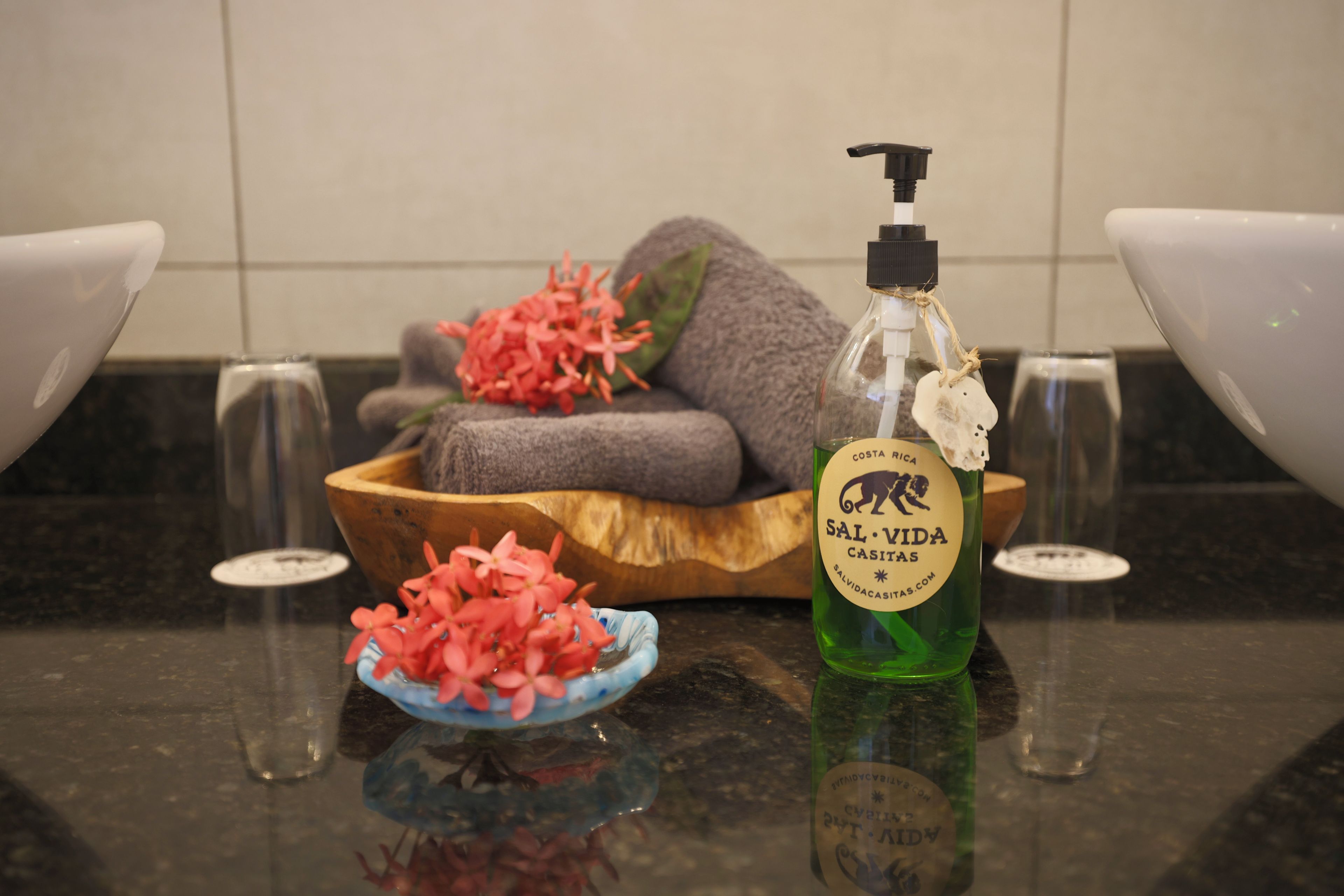 Ein Badezimmer mit einer Holtschale, die ein Handtuch und pinke Blumen enthält, einer grünen Flasche mit Pumpe und zwei weißen Waschbecken.