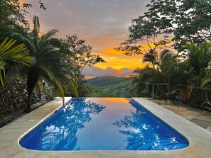 Ein Swimmingpool mit Blick auf einen Sonnenuntergang über einer hügeligen Landschaft, umgeben von tropischer Vegetation und Steinmauern.