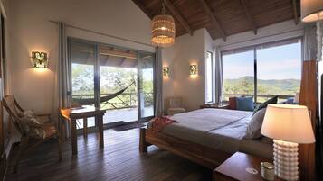 A bedroom with a large bed, wooden flooring, a hammock, and a view of the outdoors.
