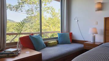 A room with a large window, a bedside table with books, a lamp, and a bed.