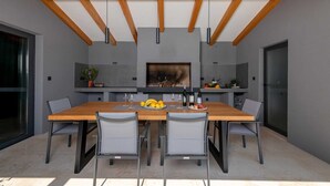 A modern dining area with a wooden table and chairs, a fireplace, and a kitchen counter with fruit and wine bottles. - Villa Marichi – Countryside Retreat w/Pool & Sauna (Marići)