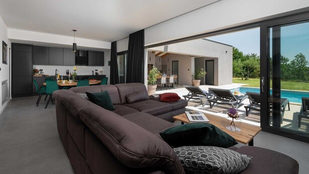 A modern living room with a large sectional sofa, a dining area, and a poolside seating area with a view of the pool. - Villa Marichi – Countryside Retreat w/Pool & Sauna (Marići)
