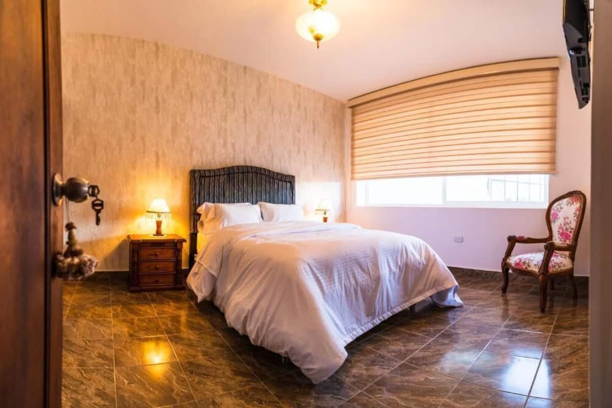 Une chambre avec un mur incurvé, un lit avec une literie blanche, une table de chevet avec une lampe et une chaise recouverte d’un tissu à motifs floraux.
