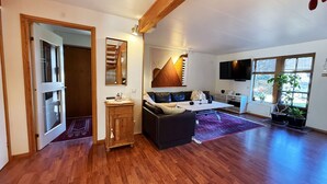 Ein geräumiges Wohnzimmer mit Holzboden, einer Couch, einem Couchtisch, einer Holzkommode und einem großen Fenster mit Pflanzen.