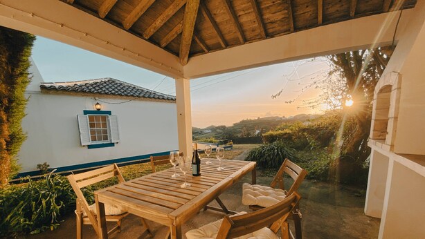 An outdoor patio with a wooden table and chairs, a bottle of wine, and glasses. There is a white building with blue trim and a window with shutters. The background features a view of hills and a sunset. - Sun Island Retreat - Great Sunny Island/Sea view (Vila do Porto)