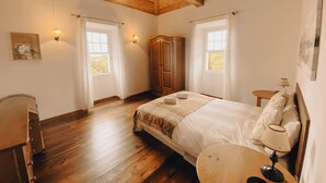 A bedroom with a wooden ceiling, two windows with curtains, a bed with a blanket, a wooden wardrobe, and a small table with a lamp. - Sun Island Retreat - Great Sunny Island/Sea view (Vila do Porto)