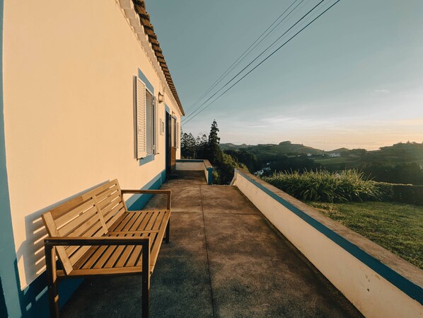 A narrow outdoor corridor with a wooden bench, a white wall with blue trim, and a view of rolling hills in the distance.