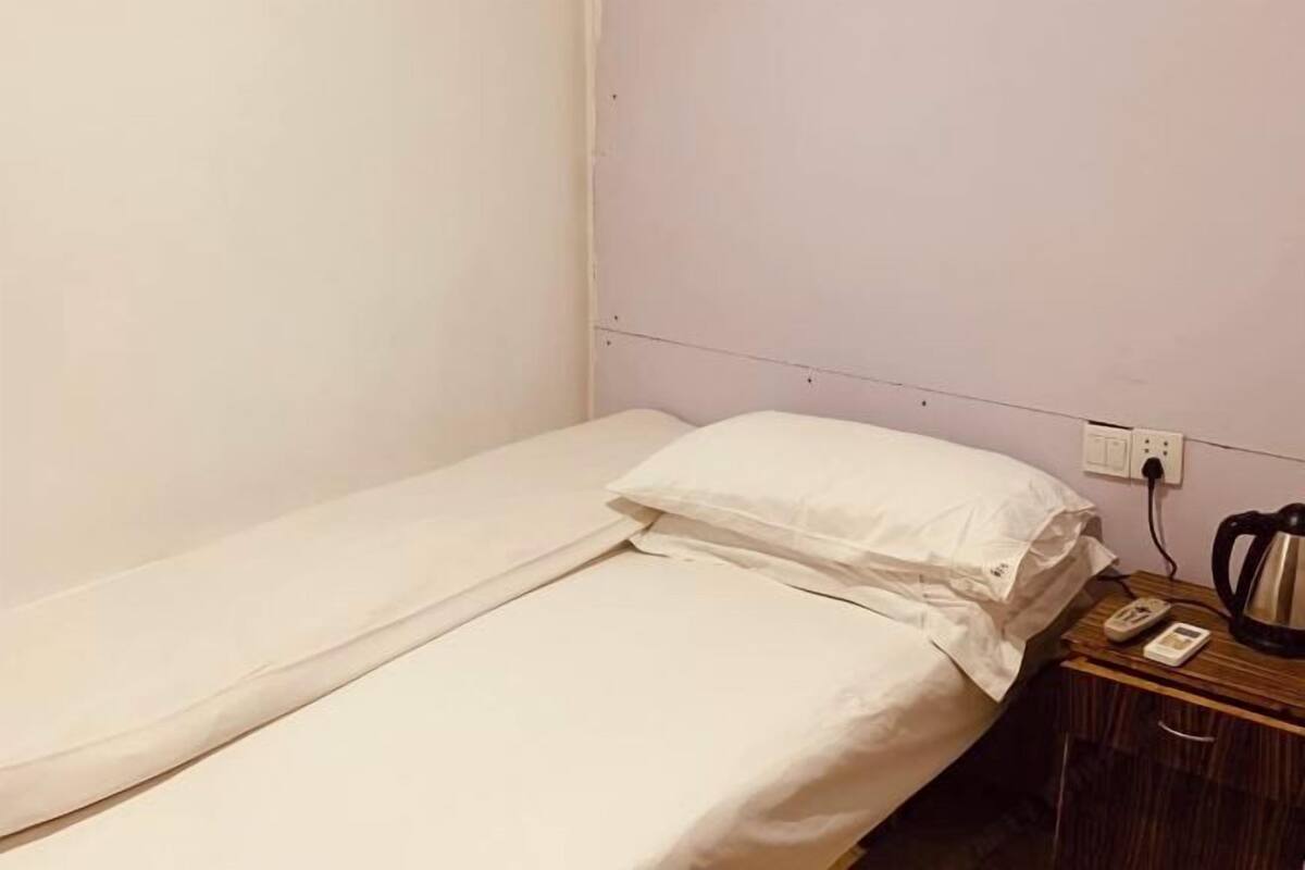 Un lit simple avec une literie blanche, une table de chevet avec une bouilloire, un téléphone et une prise électrique au mur.