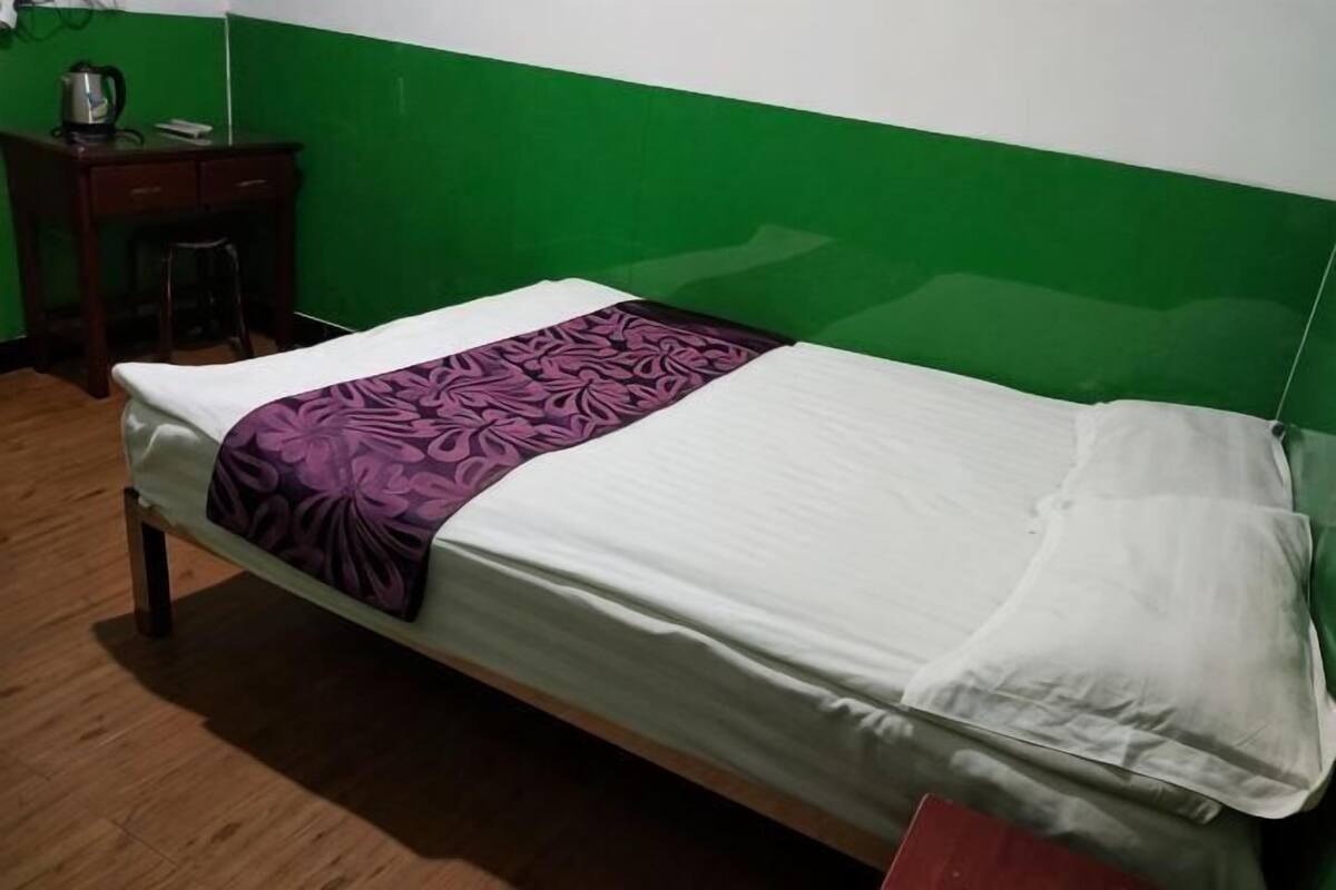 Un lit simple avec un matelas blanc et une couverture violette, un bureau en bois avec une chaise, et un mur vert.