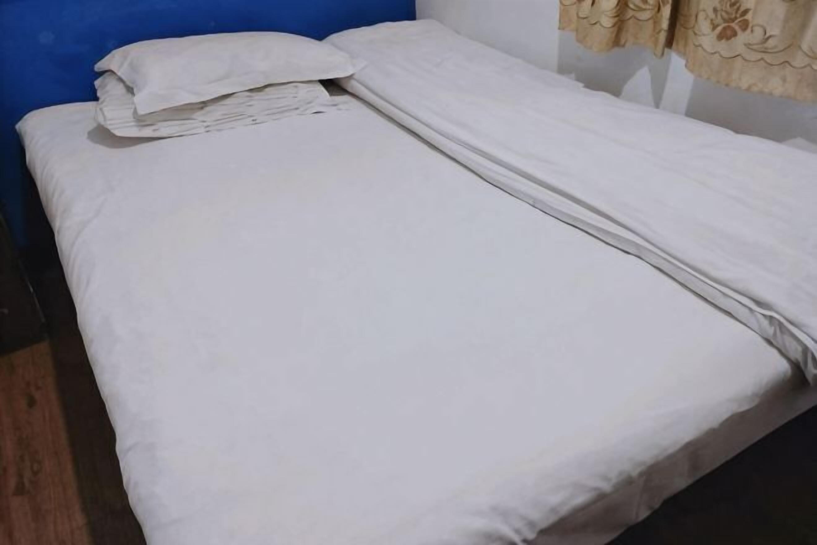Una cama individual con ropa de cama blanca, un cabecero azul y una cortina color beige.