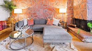 Un salon chaleureux avec un mur en pierre, un canapé d’angle gris, une table basse en verre et une cheminée.