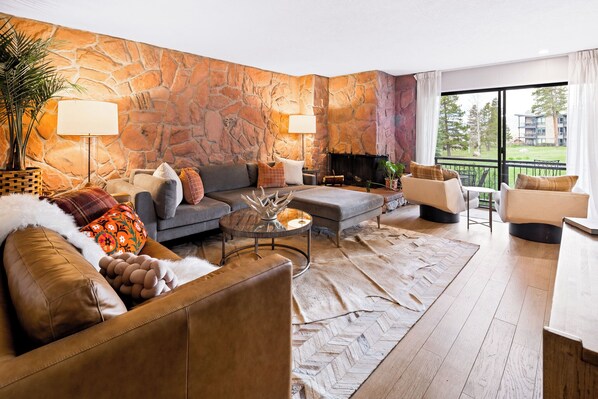 Una sala de estar moderna con una pared de piedra, sofás de cuero, una chimenea y un ventanal con vistas a árboles y edificios.