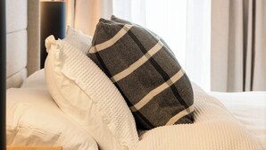 Un lit bien fait, avec une literie blanche et un coussin à carreaux, une lampe de chevet et une fenêtre avec des rideaux.