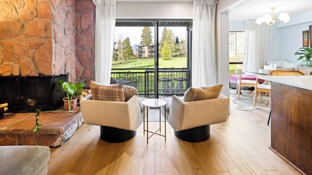 Un salon doté d’une cheminée en pierre, de deux fauteuils, d’une petite table ronde et offrant une vue sur un paysage verdoyant grâce à une porte vitrée.