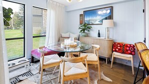 Un salon moderne avec une table à manger en verre, des chaises en bois, un canapé à motifs floraux rouges et une grande fenêtre donnant sur l’extérieur.