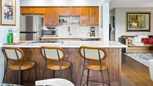 Une cuisine moderne avec des meubles en bois, un réfrigérateur en acier inoxydable, un four à micro-ondes et une machine à café. On y trouve trois tabourets de bar et une table à manger.