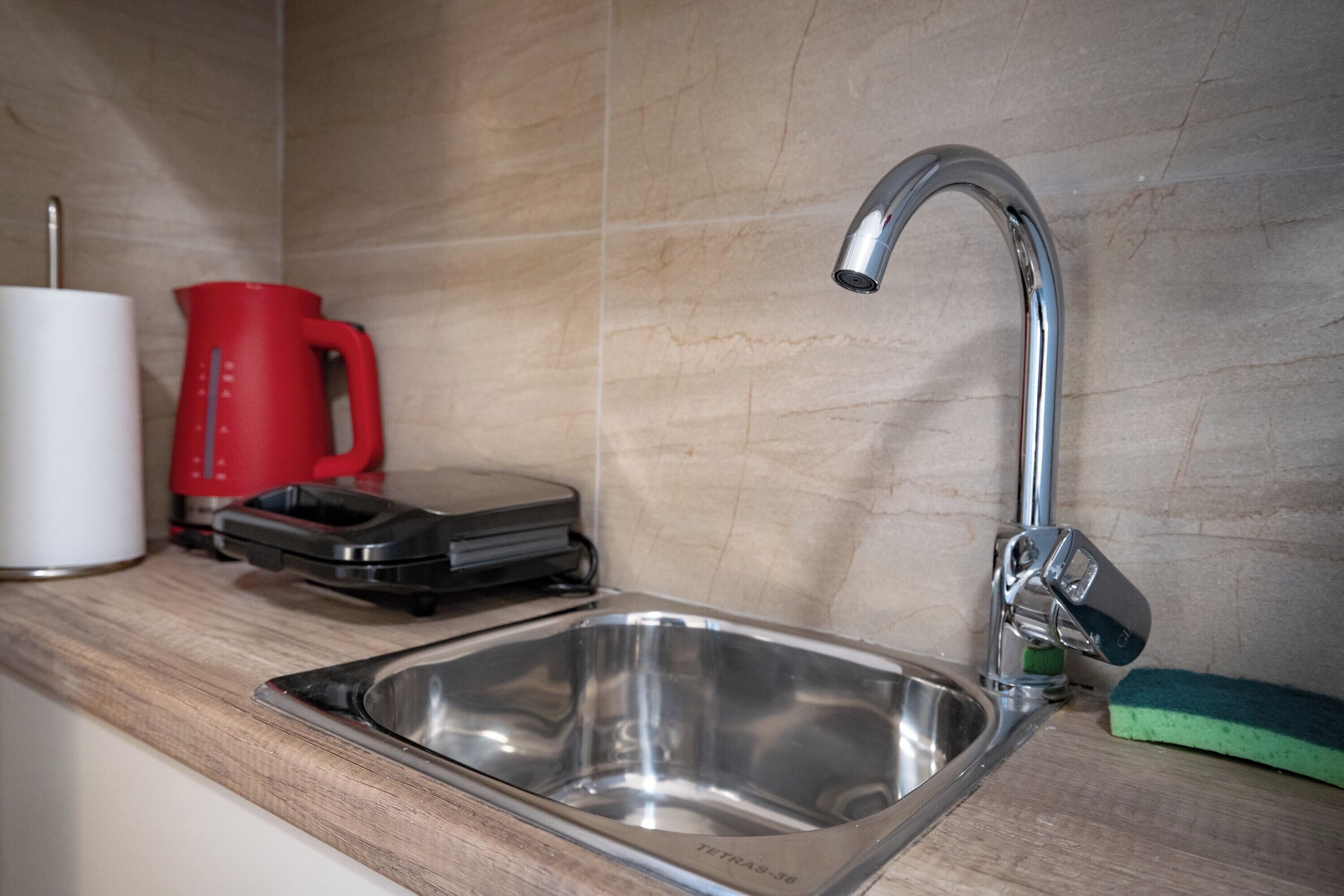 Un évier de cuisine doté d’un robinet moderne, d’une cuve en acier inoxydable, d’une cafetière rouge et d’une éponge verte posés sur le plan de travail.