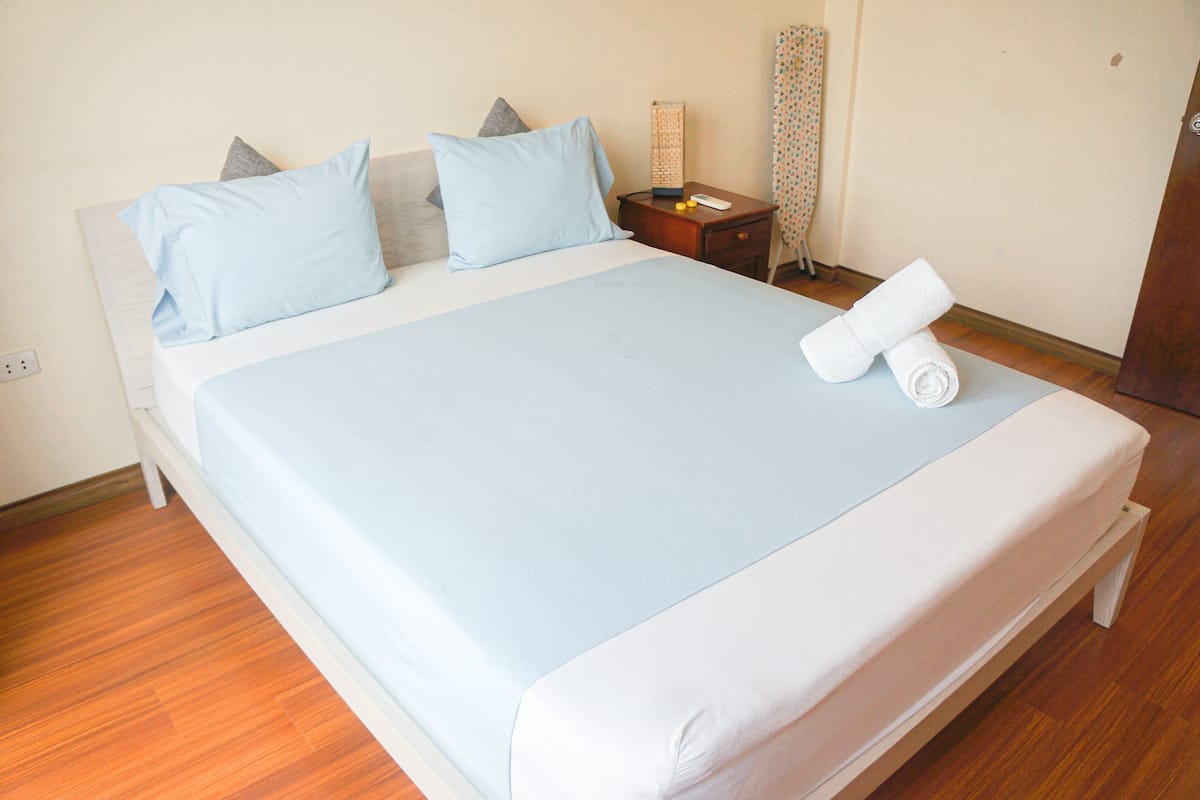Un lit bien fait, recouvert d’une couvre-lit bleu clair et agrémenté d’oreillers blancs, une table de chevet en bois avec une lampe, et une serviette blanche roulée posée sur le lit.