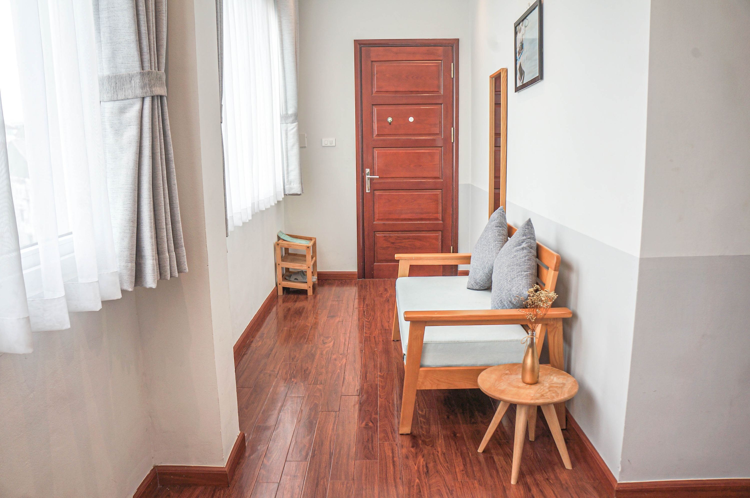 A room with a wooden floor, a bed with a grey pillow, a small wooden stool, and a door.