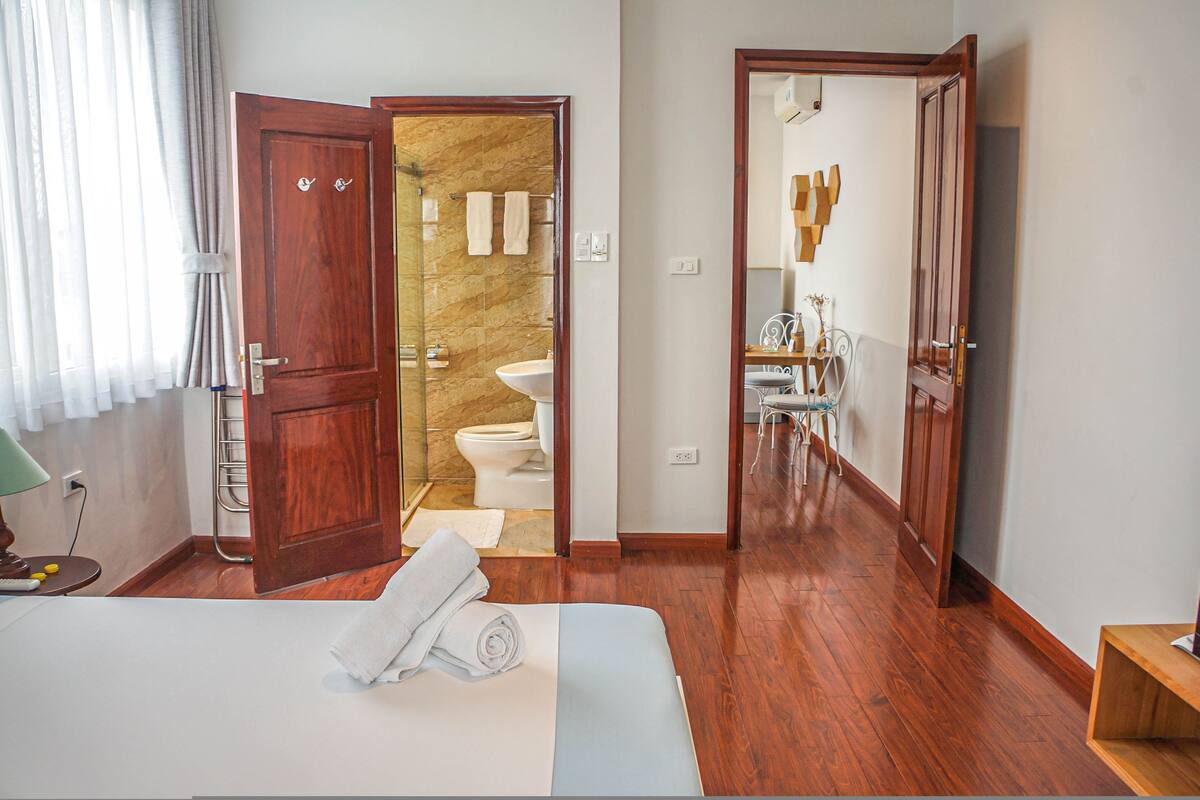 Une chambre d’hôtel avec un plancher en bois, un lit recouvert d’une courtepointe blanche et bleue, une porte en bois et une salle de bain visible à travers une porte ouverte.