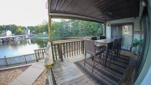 Eine Holzterrasse mit Tisch und Stühlen, mit Blick auf ein Gewässer, im Hintergrund Boote und Gebäude.