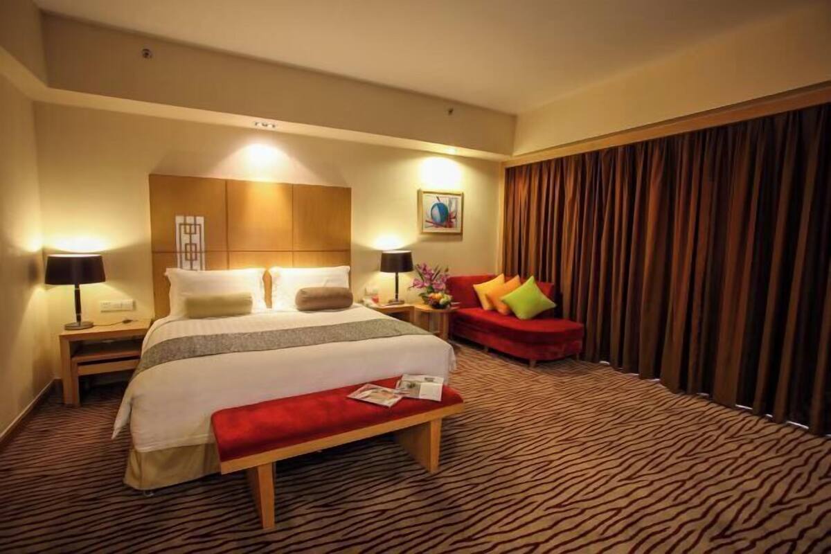 A hotel room with a large bed, a red sofa, a bench, a nightstand, a lamp, and a painting on the wall.