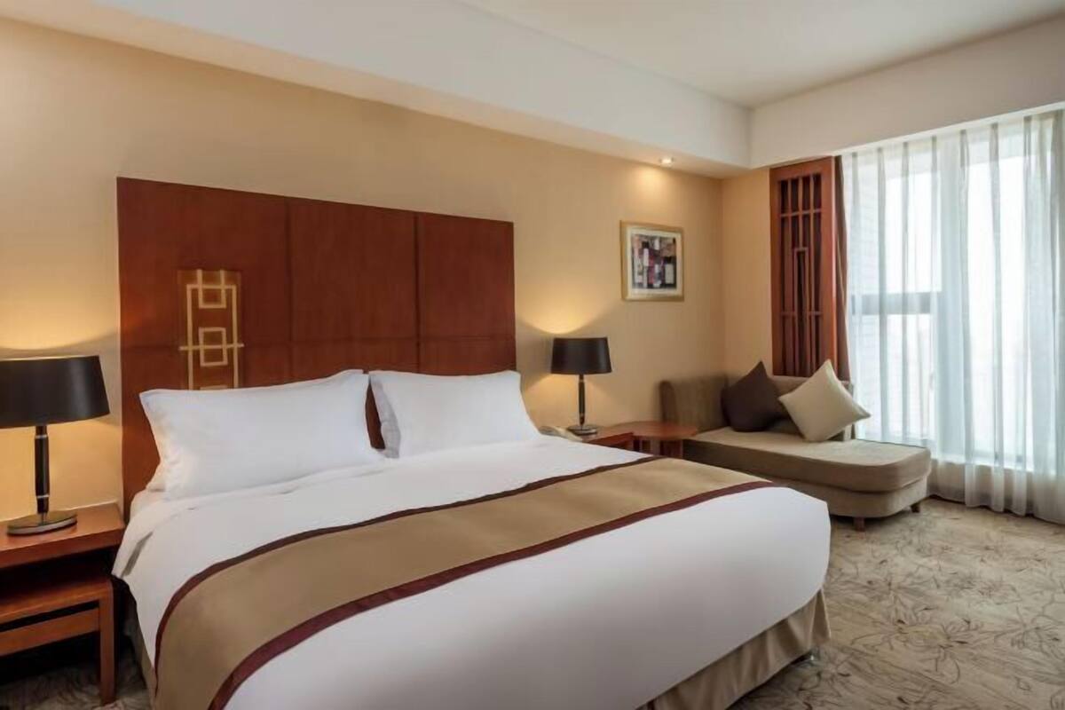 A hotel room with a large bed, a sofa, a nightstand with a lamp, and a window with curtains.