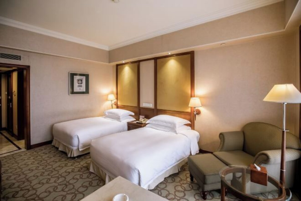 A hotel room with two beds, a chair, a table, and a lamp.