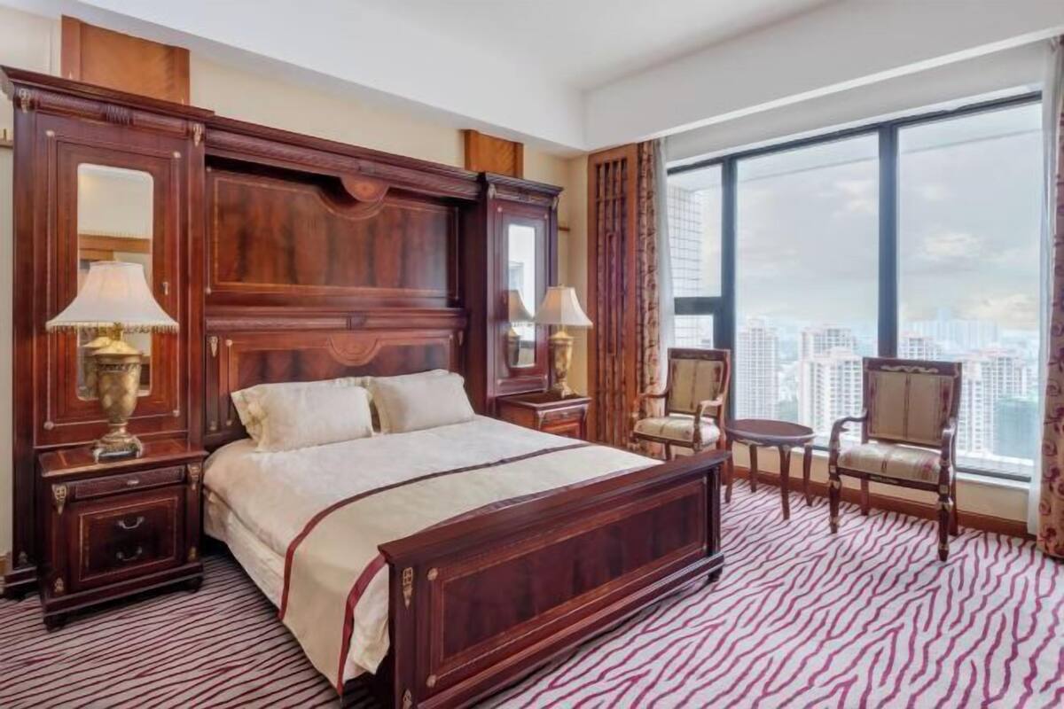 A spacious bedroom with a large bed, wooden headboard, two bedside tables with lamps, a small round table, two chairs, and a large window offering a city view.
