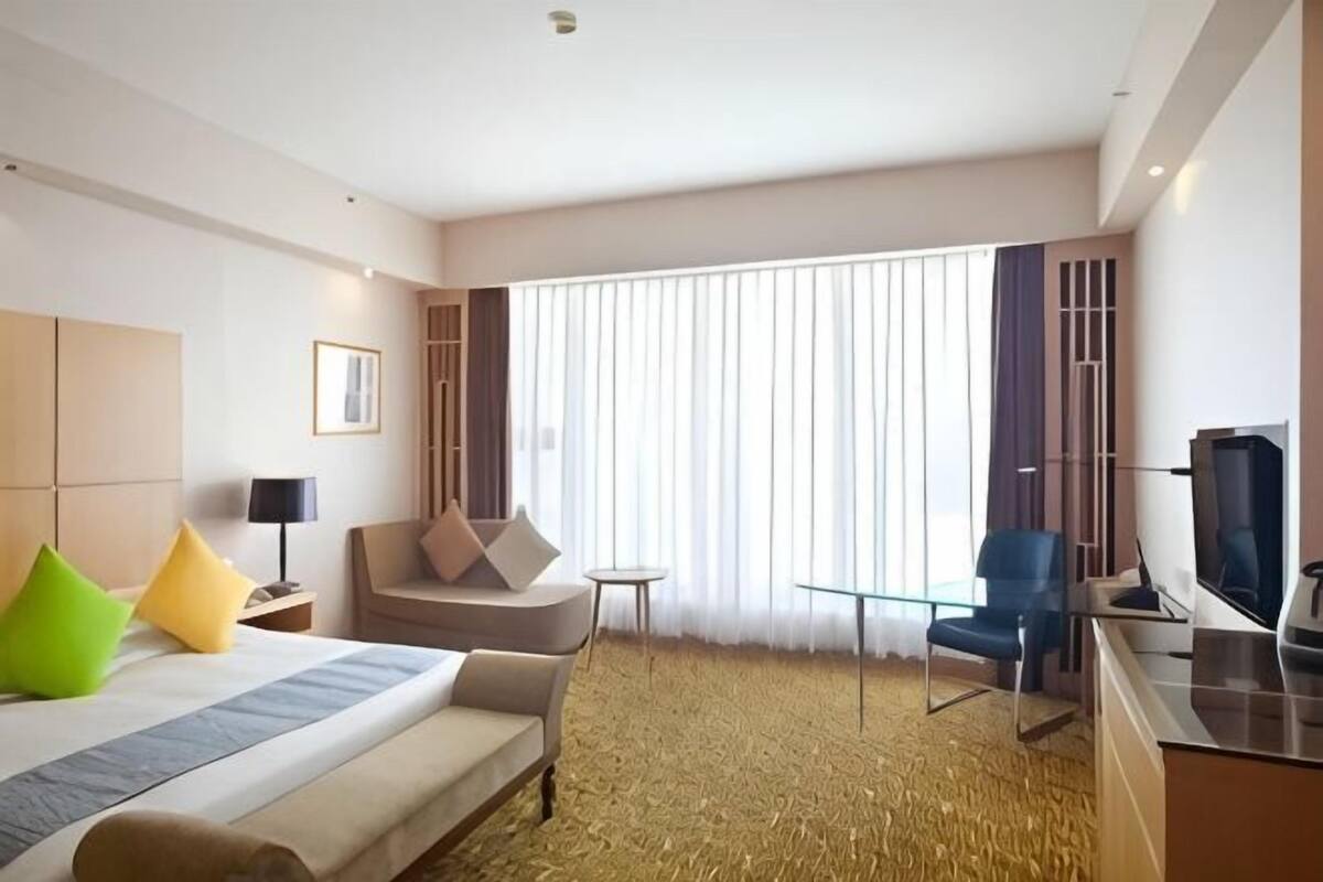 A hotel room with a bed, a desk, a chair, a TV, and a large window with curtains.