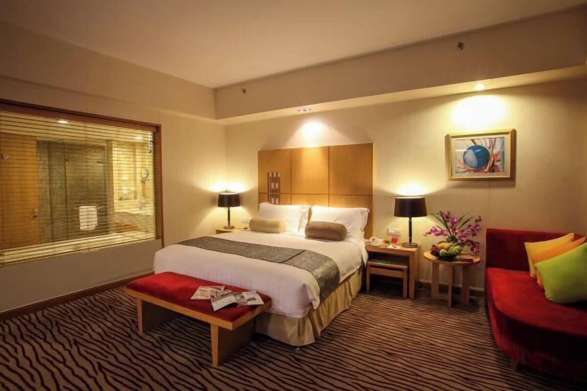 A hotel room with a large bed, bedside tables, a red sofa, a window with blinds, and a framed picture on the wall.