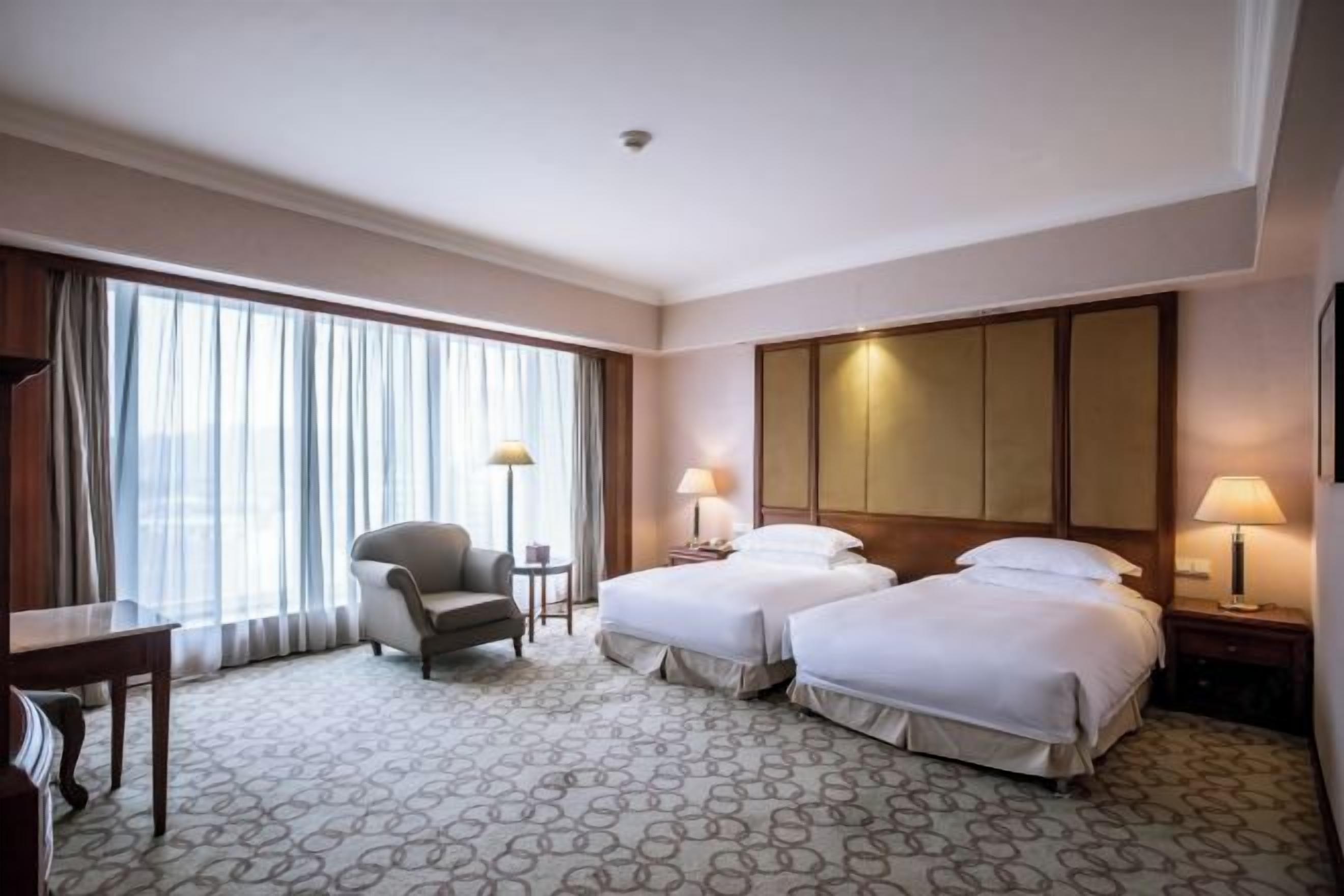 A hotel room with two beds, a chair, a desk, and a large window with curtains.