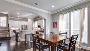 A spacious kitchen with white cabinets and a central island, a dining area with a wooden table and chairs, and large windows with curtains.