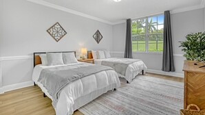 A bedroom with two beds, a window with curtains, a bedside table with a lamp, and decorative wall art.