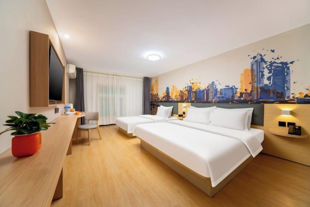 A hotel room with a bed, bedside tables, a desk, a chair, a TV, and a cityscape mural on the wall.