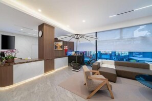 Una sala de estar moderna con un sofá, una mesa de centro y un televisor. Hay un mostrador de recepción con un reloj y una planta.