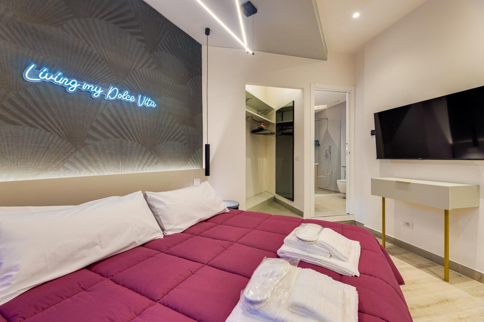 A modern hotel room with a large bed, a TV, and a neon sign on the wall.